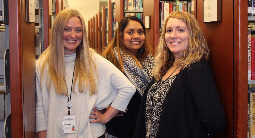 three young women stand in the aisle of the Athens Clarke County Library surrounded by shelves of books.